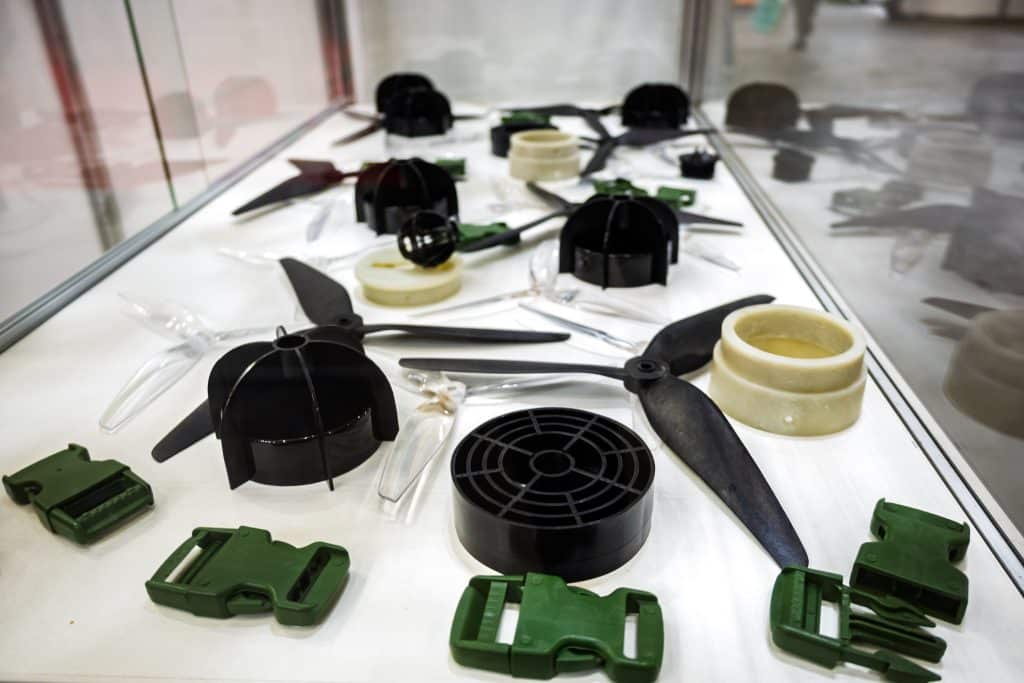 Assortment of plastic prototype parts displayed on a white surface, including drone propellers, cylinders, and green buckles, showcasing manufacturing samples or 3D printed components.